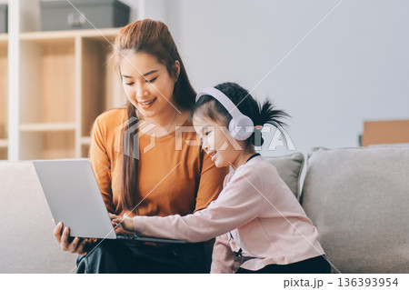 A cheerful Asian mother helps her young daughter do homework and color a book at home, while the father works on a laptop in the background. A cozy, bright, and modern family lifestyle. 136393954