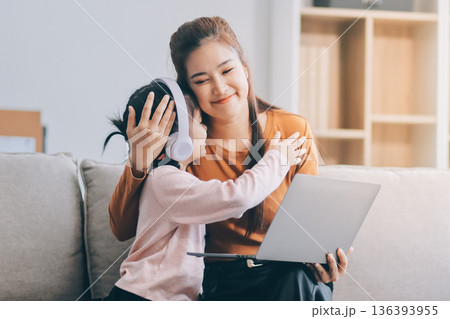 A cheerful Asian mother helps her young daughter do homework and color a book at home, while the father works on a laptop in the background. A cozy, bright, and modern family lifestyle. 136393955
