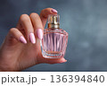 Close-up of female hand with manicured nails holding elegant pink perfume bottle isolated on gray background. Luxury fragrance, beauty elegance, cosmetic advertising and feminine scent marketing. 136394840