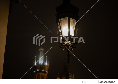 Ornate vintage street lamp illuminating a dark, misty night with a historical European tower, evoking classic urban charm and mysterious ambiance 136398002