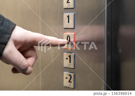 A human finger presses the illuminated 0 button on a modern stainless steel elevator control panel, symbolizing urban navigation and public building accessibility A human finger presses the illuminated 0 button on a modern stainless steel elevator control panel, symbolizing urban navigation and public building accessibility 136398014