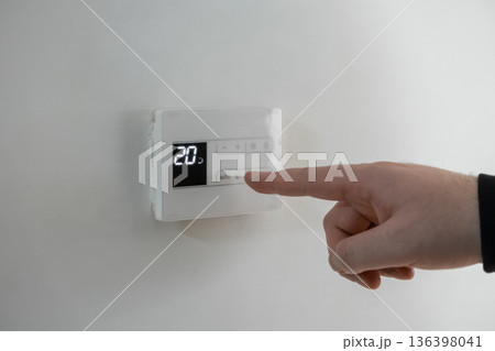 Human hand pressing the AUTO button on a white digital thermostat, displaying 20 degrees for comfortable home climate control and efficient energy management 136398041