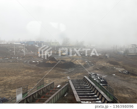 Industrial construction site with heavy equipment, temporary buildings, and distant railroad tracks, obscured by dense fog, representing infrastructure progress Industrial construction site with heavy equipment, temporary buildings, and distant railroad tracks, obscured by dense fog, representing infrastructure progress 136398096