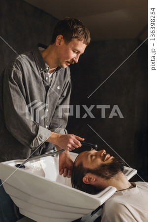 Barber washing client hair with shampoo at salon sink. Professional male grooming service, relaxing head wash process, close up of hairstyling care in modern barbershop. Barber washing client hair with shampoo at salon sink. Professional male grooming service, relaxing head wash process, close up of hairstyling care in modern barbershop. 136398284
