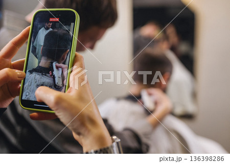 Smartphone recording barber haircut process in modern barbershop. Close up of mobile phone screen filming professional hair trimming, male grooming, content creation, social media concept. Smartphone recording barber haircut process in modern barbershop. Close up of mobile phone screen filming professional hair trimming, male grooming, content creation, social media concept. 136398286