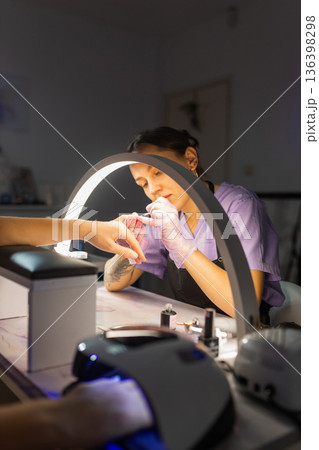 Professional nail technician performing manicure for female client under lamp in beauty salon. Nail care service, cosmetic treatment, hygiene, modern manicure workspace concept. Professional nail technician performing manicure for female client under lamp in beauty salon. Nail care service, cosmetic treatment, hygiene, modern manicure workspace concept. 136398298