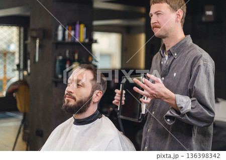 Barber showing haircut result to male client using mirror in barbershop. Professional consultation, beard and hair styling, men grooming service, modern barber salon interior. 136398342