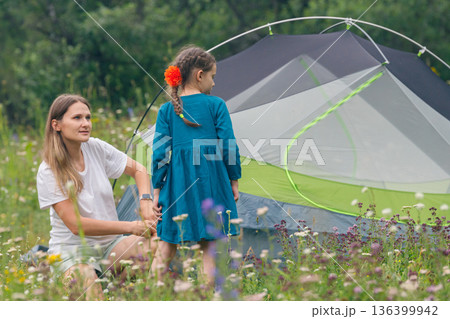 A mother and her daughter are joyfully enjoying a delightful camping experience in a beautiful blooming meadow 136399942