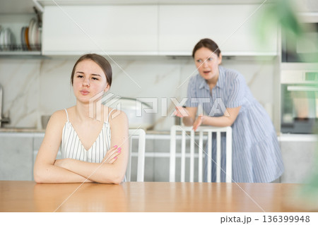 Offended young girl sitting at the kitchen table, mother standing behind 136399948