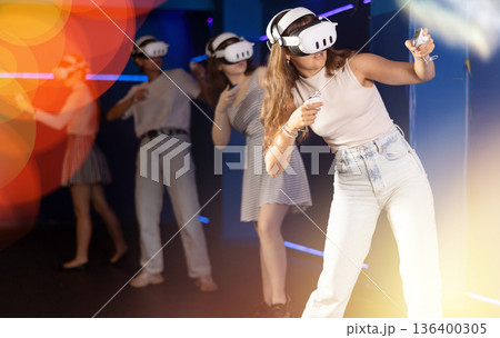 Emotional girls in VR headsets fully immersed in game, engaged in virtual reality world, manipulating objects or shooting targets with joysticks in hands 136400305