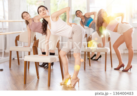 Leaning on chair seat with hand foot, young woman performs lap dance during group workout. 136400483