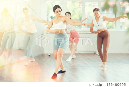 Young female ballet dancer executing dance moves with focused precision among group of people rehearsing in ballet studio Young female ballet dancer executing dance moves with focused precision among group of people rehearsing in ballet studio 136400496