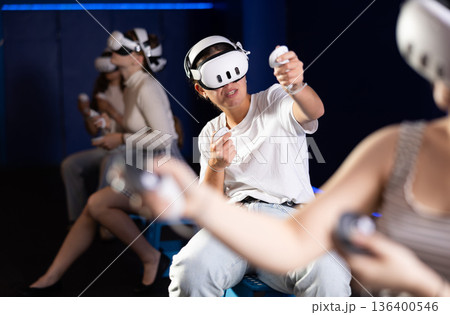 Portrait of young girlfriends using virtual reality goggles controlling joysticks sitting in room with friends 136400546