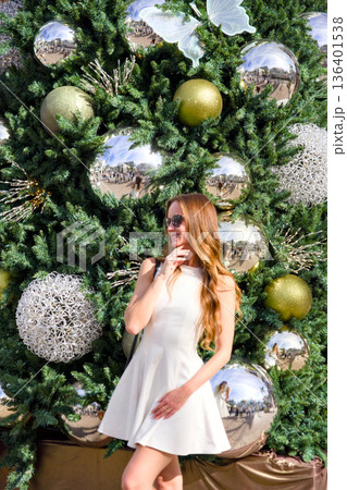 Woman in white dress posing near decorated christmas tree outdoors 136401538