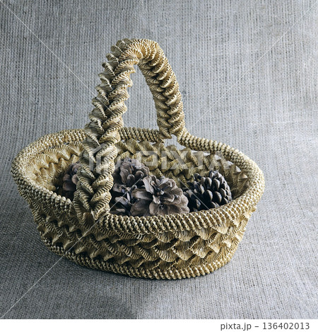 A woven straw basket against a burlap background. Close-up. Eco-friendly. The handmade basket is filled with pine cones 136402013