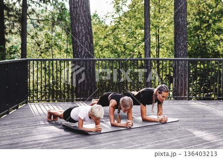 Active mother exercising together with children while holding plank pose on yoga mats outdoors. Family fitness and healthy lifestyle, strength training, summer workout on wooden terrace in nature Active mother exercising together with children while holding plank pose on yoga mats outdoors. Family fitness and healthy lifestyle, strength training, summer workout on wooden terrace in nature 136403213