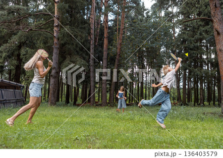 Active family enjoying outdoor leisure together, playing badminton on grass meadow with pine trees. Mother and children having fun in nature, summer weekend, wide angle full length 136403579