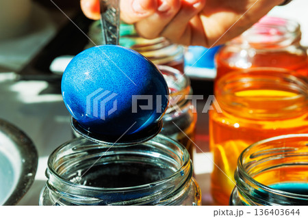 Close up of a hand lifting a blue dyed easter egg on a spoon above a glass jar, with orange dye jars glowing in sunlight behind. Close up of a hand lifting a blue dyed easter egg on a spoon above a glass jar, with orange dye jars glowing in sunlight behind. 136403644