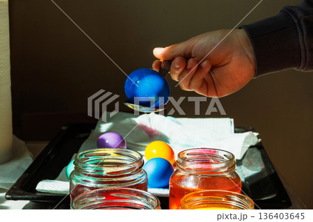 Sunlit close up of a hand holding a blue dyed easter egg on a spoon above glass food dye jars, with other colored eggs drying on a tray 136403645