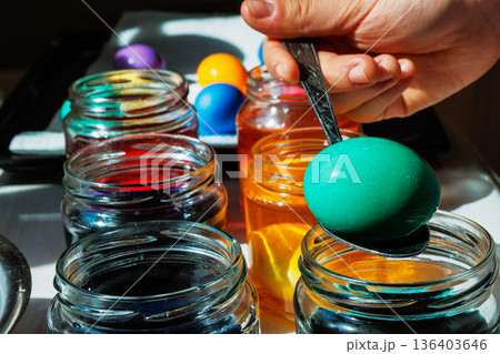 Close up of a hand lifting a green dyed easter egg on a spoon above a glass jar, with orange dye jars glowing in sunlight behind. 136403646