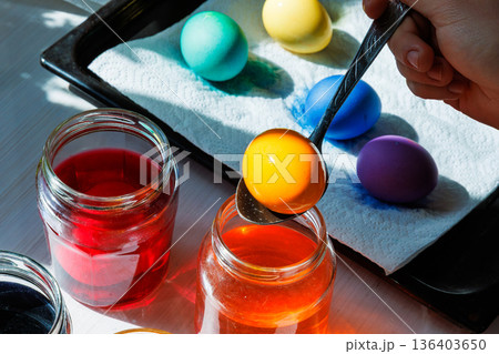 Close up of the spoon lifting orange dyed egg above jar with colorful easter eggs on tray 136403650