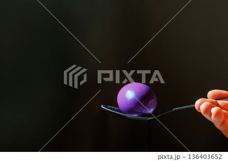 Hand holding a spoon with a purple dyed easter egg with copy space against black background 136403652