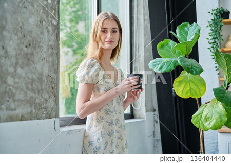 Young beautiful smiling romantic girl looking at camera with cup, standing near window 136404440