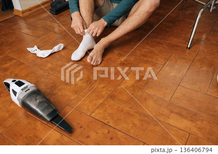 Close-up cropped shot of man sitting on wooden floor removing white sock, holding leg, with a handheld vacuum cleaner and upright vacuum in background, representing domestic routine and self-care. Close-up cropped shot of man sitting on wooden floor removing white sock, holding leg, with a handheld vacuum cleaner and upright vacuum in background, representing domestic routine and self-care. 136406794