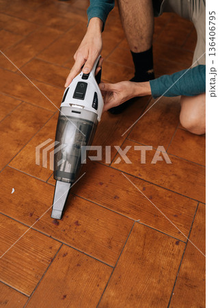 Vertical shot of person cleaning up dirt and debris from wooden floor using compact cordless vacuum, effectively maintaining cleanliness and hygiene in living room. Concept of household chores. 136406795