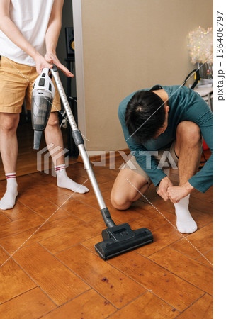 Vertical shot of two men performing household maintenance together, with one using vacuum to tidy up wooden flooring, another prepares for activity, emphasizing teamwork and domestic responsibility. Vertical shot of two men performing household maintenance together, with one using vacuum to tidy up wooden flooring, another prepares for activity, emphasizing teamwork and domestic responsibility. 136406797