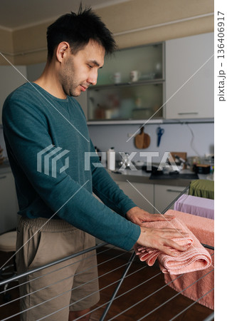 Vertical shot of man performing daily household chores, hanging freshly washed towels on drying rack in kitchen, demonstrating domestic responsibility. Concept of household chores and housework. Vertical shot of man performing daily household chores, hanging freshly washed towels on drying rack in kitchen, demonstrating domestic responsibility. Concept of household chores and housework. 136406917