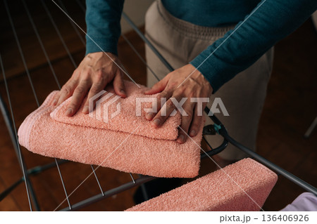 Person folding soft pink towels on drying rack, organizing clean laundry indoors, routine household chore showing careful hands, neat fabric, everyday home care, close-up cropped shot. 136406926