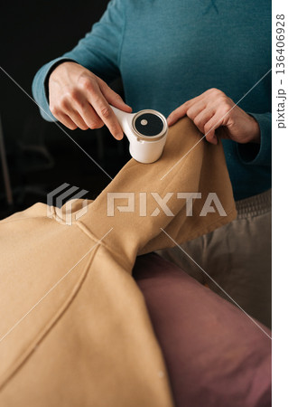 Vertical cropped shot of housekeeper using electric fabric shaver removing lint from beige sweatshirt sleeve for garment care and renewed clothing appearance, maintaining textile quality. Vertical cropped shot of housekeeper using electric fabric shaver removing lint from beige sweatshirt sleeve for garment care and renewed clothing appearance, maintaining textile quality. 136406928