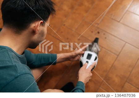 Rear top view of man cleaning wooden floor with portable handheld vacuum cleaner, tidying crumbs and dust, maintaining household hygiene and spotless home environment. 136407036