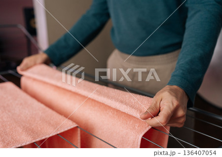 Cropped shot of male hands smoothing clean, damp orange towel over metal bars of indoor drying rack, completing everyday laundry chore in bright, organized home setting. 136407054