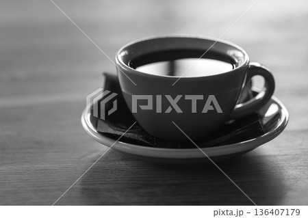 A monochrome image of a simple coffee cup on a saucer, set on a wooden table 136407179