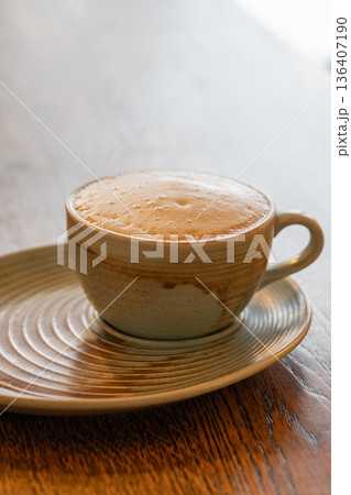 A warm ceramic coffee cup with foam sits on a matching saucer atop a wooden table 136407190