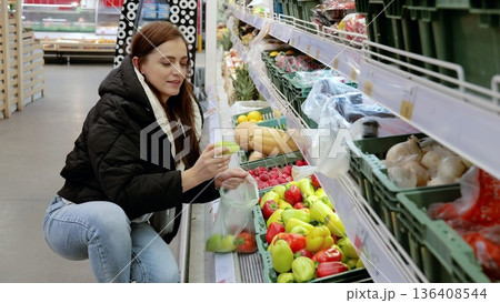 Young lady carefully picking out fresh bell peppers in a brightly lit supermarket produce section, and placing them in a reusable bag 136408544