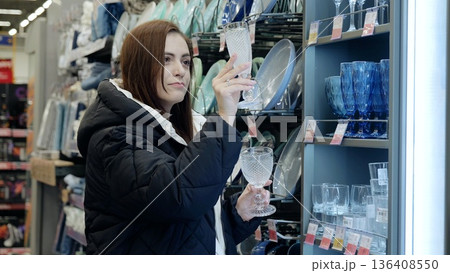Young woman carefully selecting crystal glasses in the houseware department of a supermarket before making a purchase Young woman carefully selecting crystal glasses in the houseware department of a supermarket before making a purchase 136408550