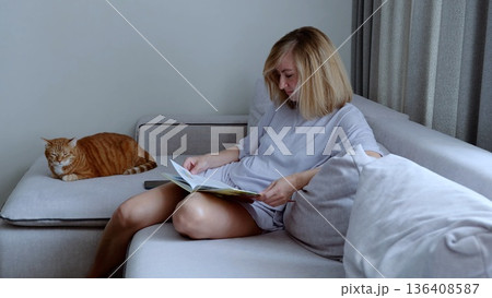 Tranquil domestic scene, woman reading with her ginger cat peacefully sleeping beside her on a comfortable sofa in a cozy living room 136408587