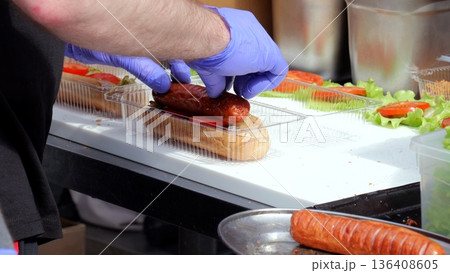 Food vendor preparing, Vendor assembling sausages at food festival, Closeup of hot dog preparation at lively outdoor event, Chef with gloves preparing sausage sandwiches amidst bustling food market Food vendor preparing, Vendor assembling sausages at food festival, Closeup of hot dog preparation at lively outdoor event, Chef with gloves preparing sausage sandwiches amidst bustling food market 136408605