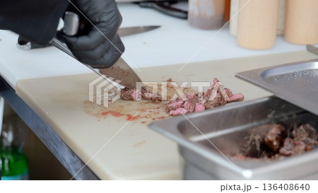 Hands slicing smoked brisket on board, vendor wearing black gloves carves tender brisket into thin slices on yellow board beside stainless tray, smoky aroma, casual outdoor festival catering scene Hands slicing smoked brisket on board, vendor wearing black gloves carves tender brisket into thin slices on yellow board beside stainless tray, smoky aroma, casual outdoor festival catering scene 136408640