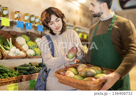 Young girl choosing mango from basket held by male seller grocery market Young girl choosing mango from basket held by male seller grocery market 136408732