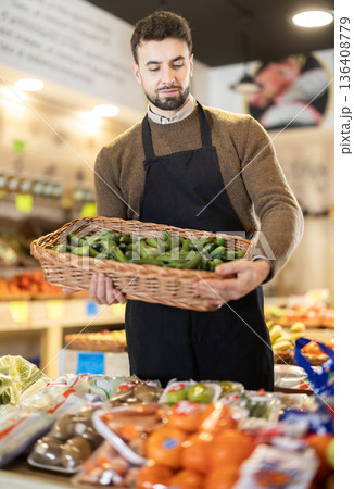 Young male seller holding basket of green long peppers in grocery market Young male seller holding basket of green long peppers in grocery market 136408779
