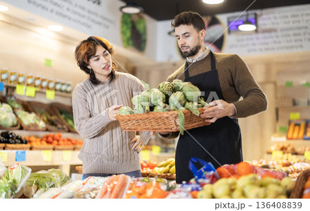 Young girl choosing artichoke from basket held by male seller grocery market Young girl choosing artichoke from basket held by male seller grocery market 136408839