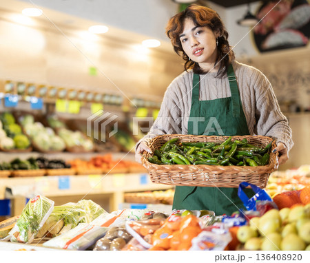 Woman employees in uniform carries wicker basket with green peppers in grocery shop Woman employees in uniform carries wicker basket with green peppers in grocery shop 136408920