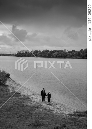 Father and daughter holding hands on river shore under cloudy sky, black and white 136409299