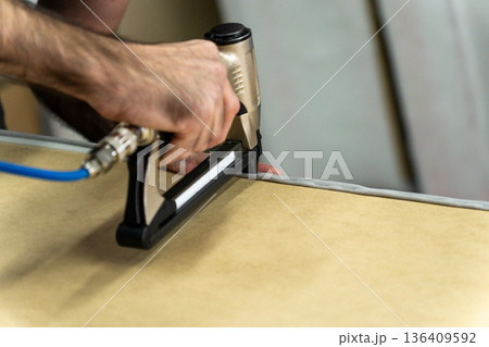 Close-up of upholsterer using a pneumatic staple gun to attach fabric to a wooden furniture frame. Close-up of upholsterer using a pneumatic staple gun to attach fabric to a wooden furniture frame. 136409592