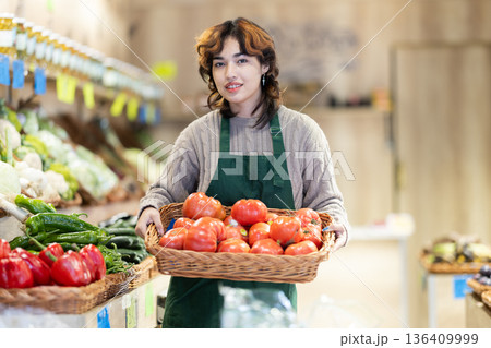 Girl employees in uniform carries a wicker basket with tomatoes in grocery shop Girl employees in uniform carries a wicker basket with tomatoes in grocery shop 136409999