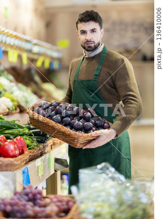 Man employees in uniform carries wicker basket with plums in grocery shop Man employees in uniform carries wicker basket with plums in grocery shop 136410006